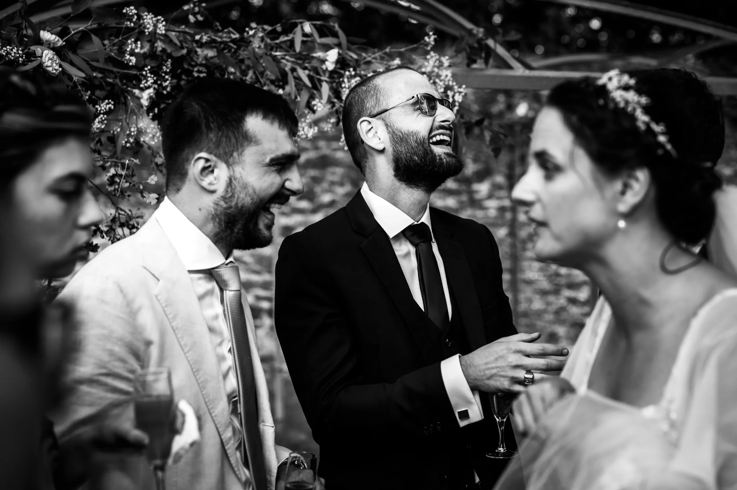 lors du cocktail d'un mariage les invités rigolent en seond plan et en premier plan on voit la mariée en grand discussion, cette photo est en noir et blanc